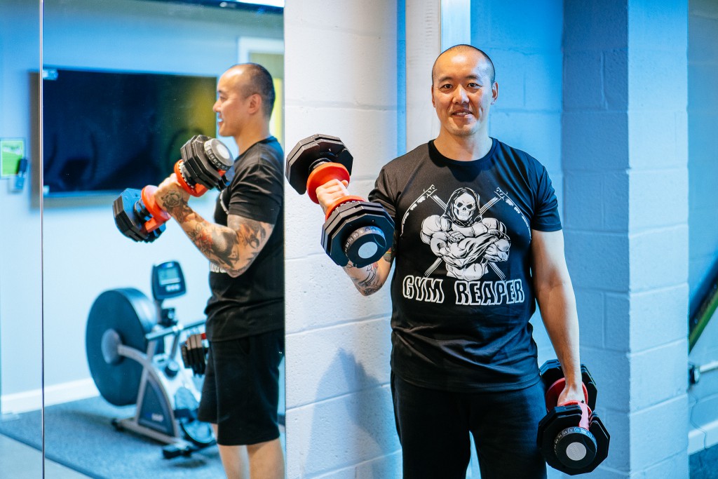kit wong of unwonted apparel stands in a gym holding large dumbells in both hands